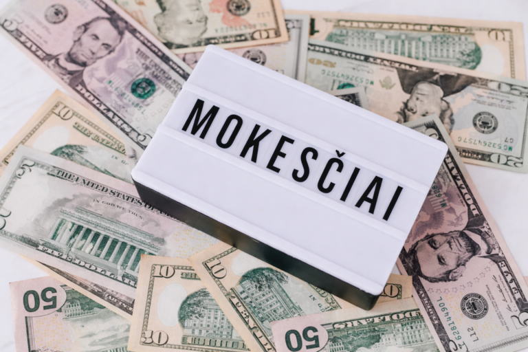 Nuotrauka, kurioje švieslentė su užrašu „MOKESČIAI“ padėta ant JAV dolerių banknotų.