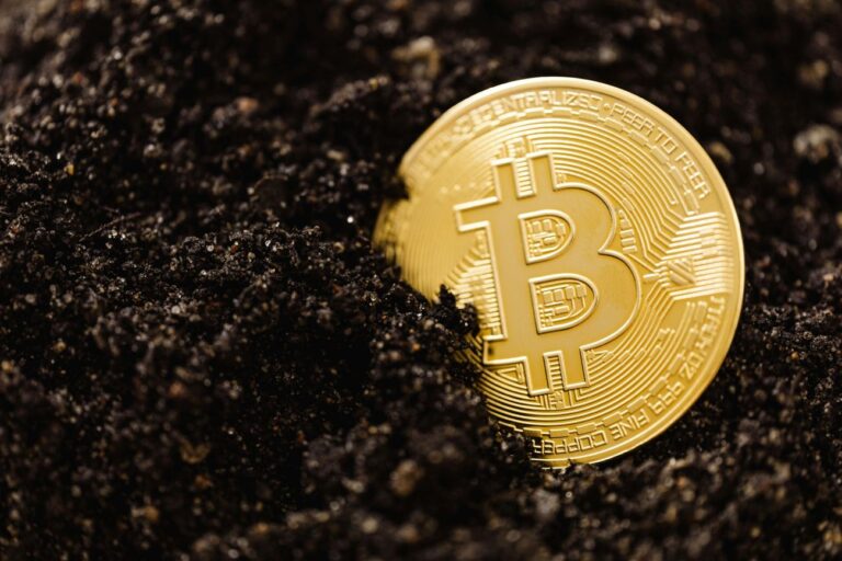 Stambiu planu pavaizduota auksinė „Bitcoin“ moneta, iš dalies įkasta tamsioje žemėje. Nuotrauka simbolizuoja kriptovaliutų kasimą ir pastangas išgauti skaitmeninę vertę.