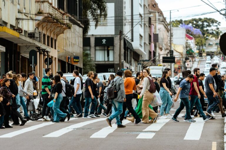Miesto gatvėje per perėją einanti žmonių minia, iliustruojanti rinkos nuotaikos, minios elgsenos ir „contrarian“ investavimo strategijos temą.
