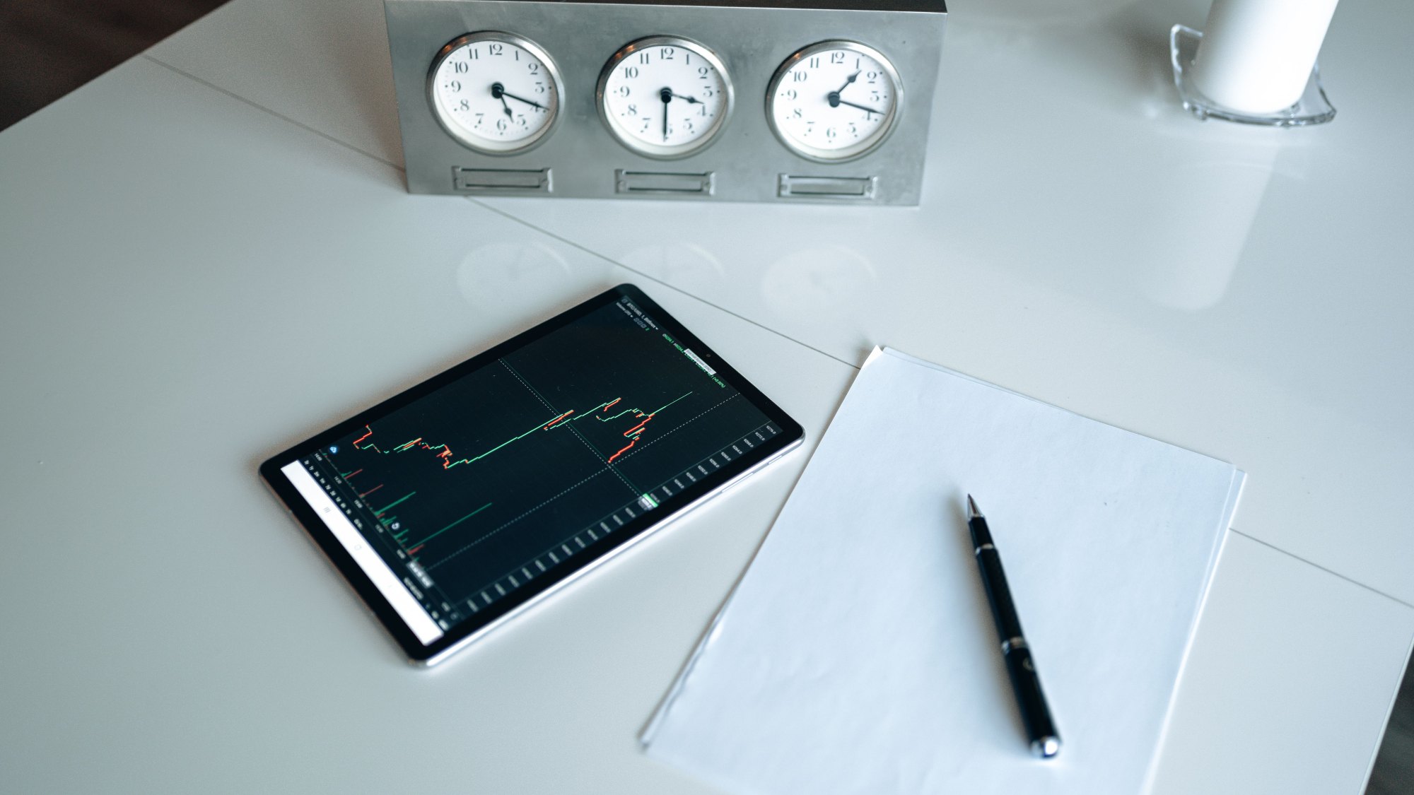 Ant stalo padėtas planšetinis kompiuteris su akcijų kainos grafiku, šalia matomi laikrodžiai, užrašinė ir rašiklis, iliustruojantys „market timing“ strategiją ir bandymą pasirinkti tinkamą laiką pirkti akcijas.