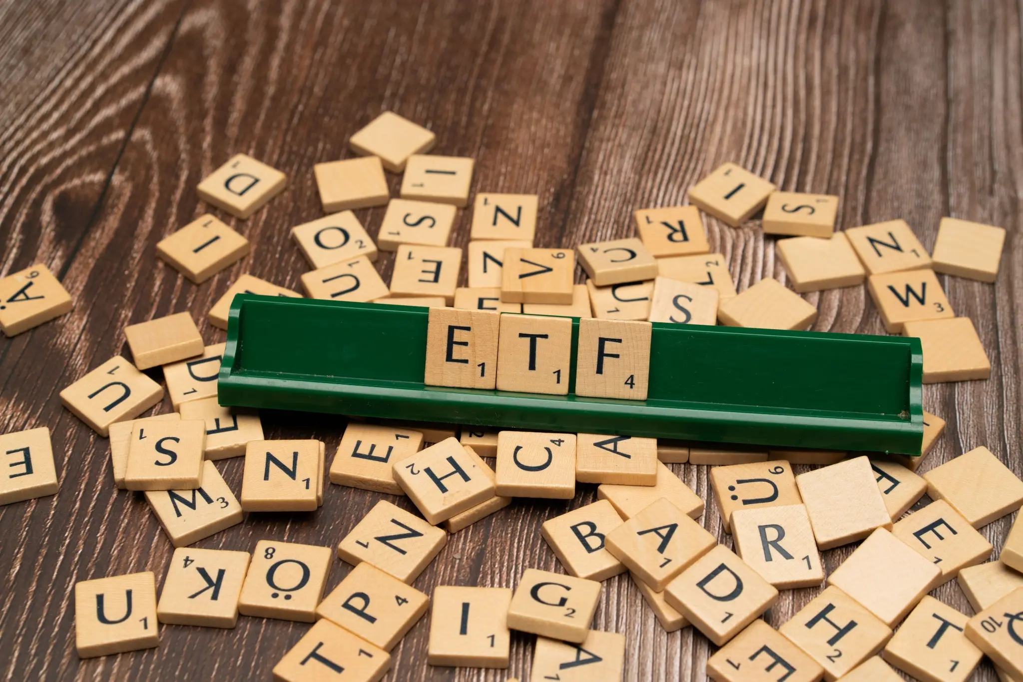 Medinių raidžių kaladėlės išdėliotos ant medinio paviršiaus, o centre ant žalio laikiklio sudėliotas žodis „ETF“. Vaizdas iliustruoja ETF temą.